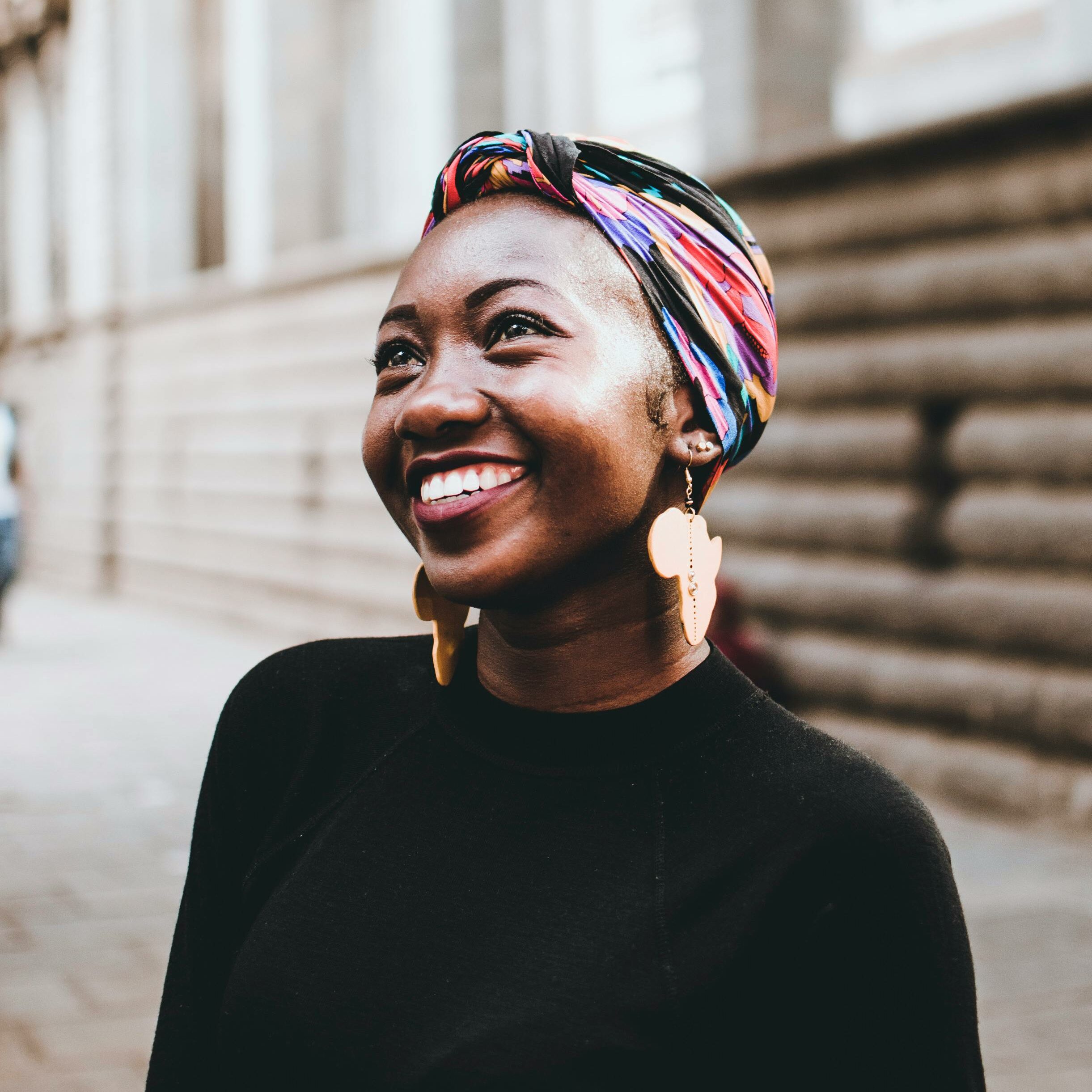 Smiling woman wearing vibrant headscarf and earrings, captured in an urban street setting.